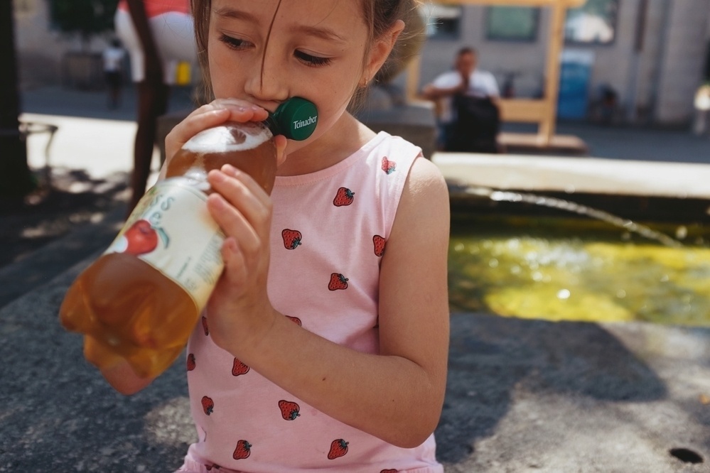 Young girl drinks juice from a bottle while having a break from playing with water at a public city fountain on a hot summer day