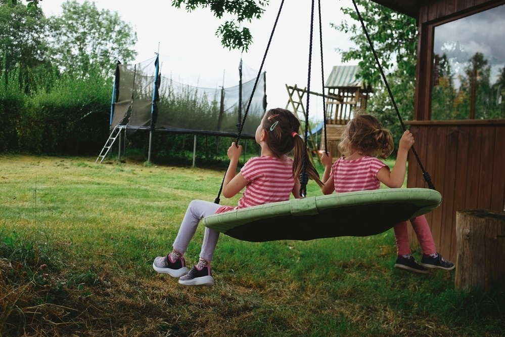 Two children playing on a nest swing next to a wooden hut, and a trampoline in the background