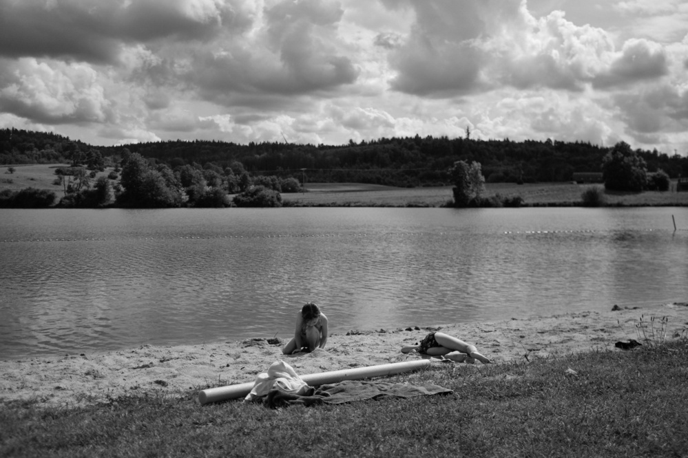 Black-and-white photo of a lake with a small sand beach where one child is playing and another one is sunbathing. In the background there’s hills covered in trees and a cloudy sky.