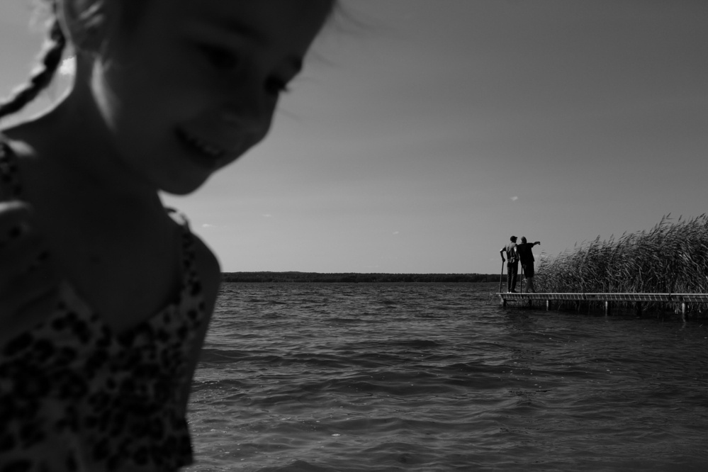 A blurred child in the foreground with a background featuring two people on a pier by a lake with reeds.