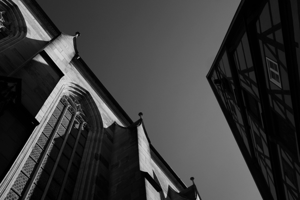 Black-and-white contrast-rich photo of the top of two buildings: on the left side, a gothic cathedral, on the right a half-timbered house, in the background the clear sky.