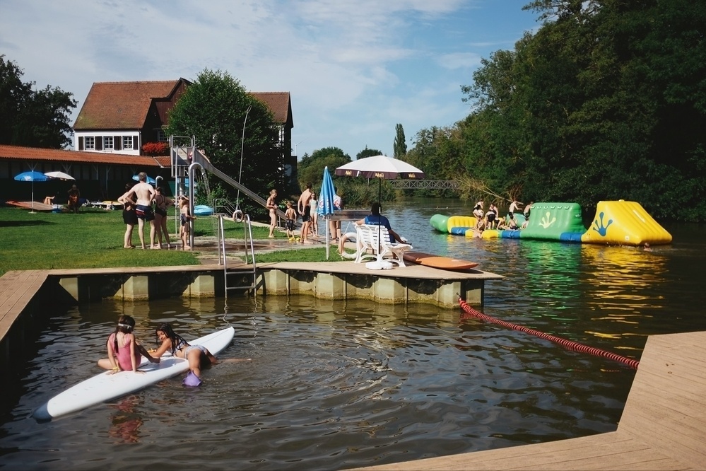 Overview of the Wörnitzstandbad river beach. On the left side, some people hang around on the lawn. In the foreground, two children lie on a surfboard in the water. On the right side, a large inflatable floats on the river and several teenagers can be seen playing.