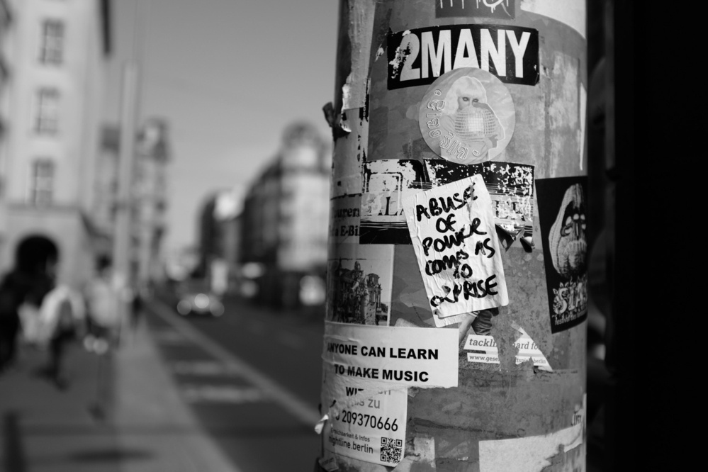 Close-up of a pole covered in various stickers and posters, with one sticker reading “ABUSE OF POWER COMES AS NO SURPRISE” in focus.