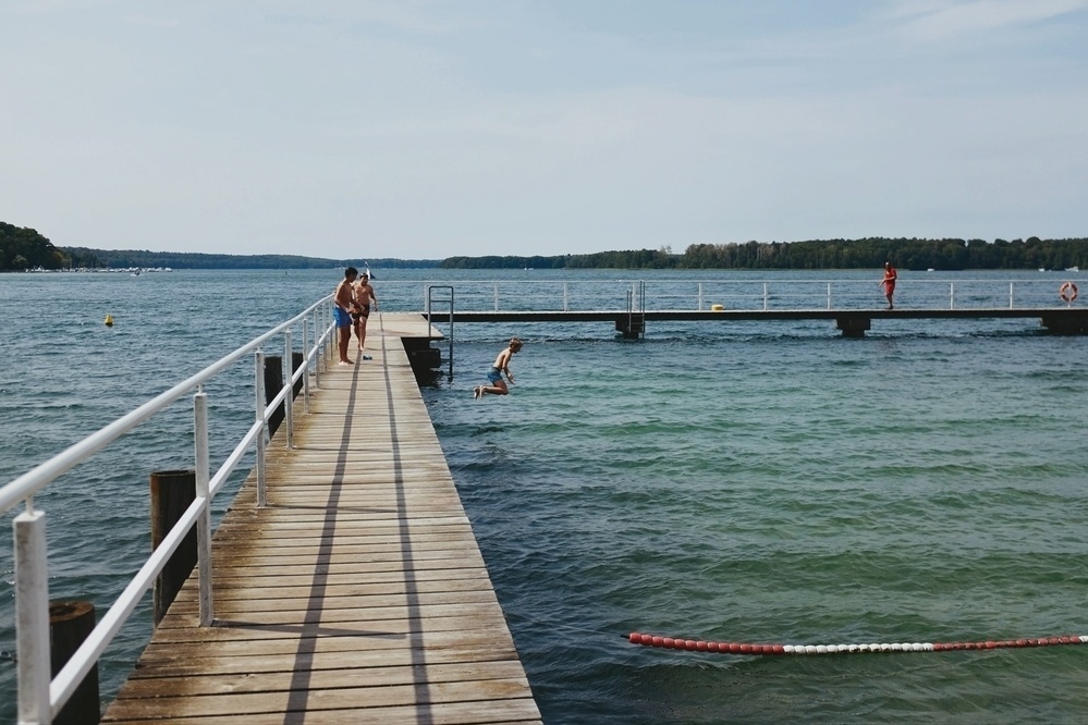 Wooden pier extending into a lake with people enjoying the water, one child jumping off the side, surrounded by forested land.