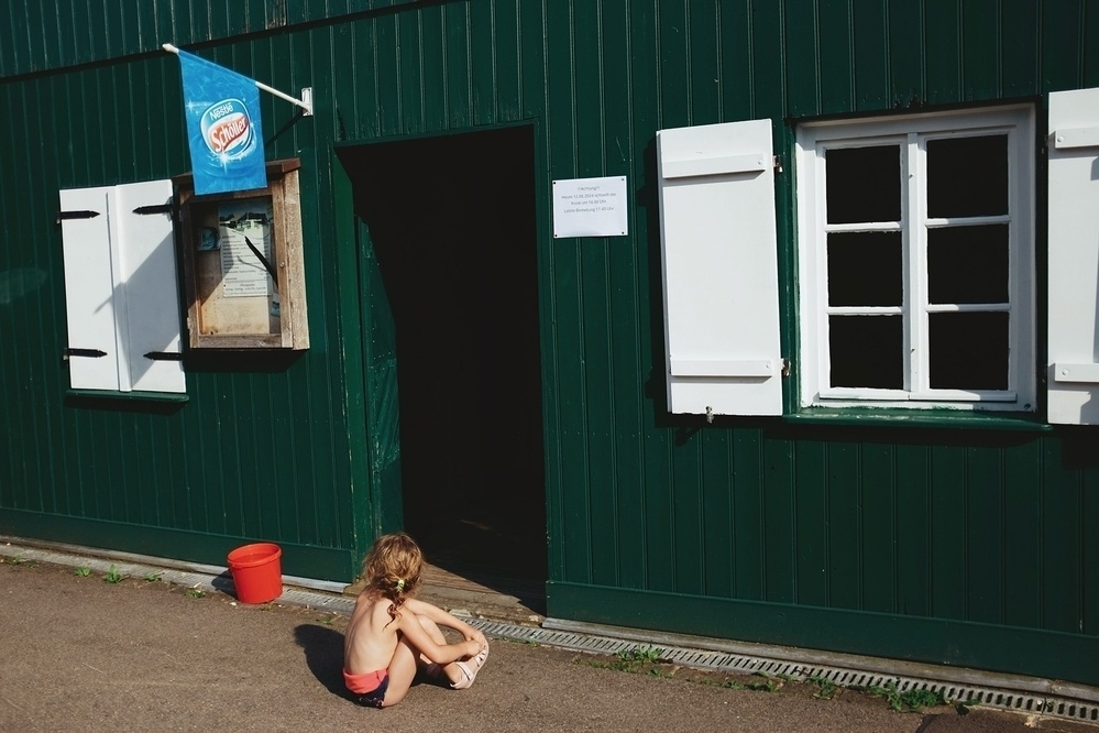 Young child sits on the ground while putting on shoes in front of the entrance door to a green wooden beach kiosk.