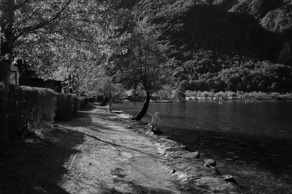 Black-and-white photo of a dirt pathway alongside Lake Lugano with a child playing under a tree