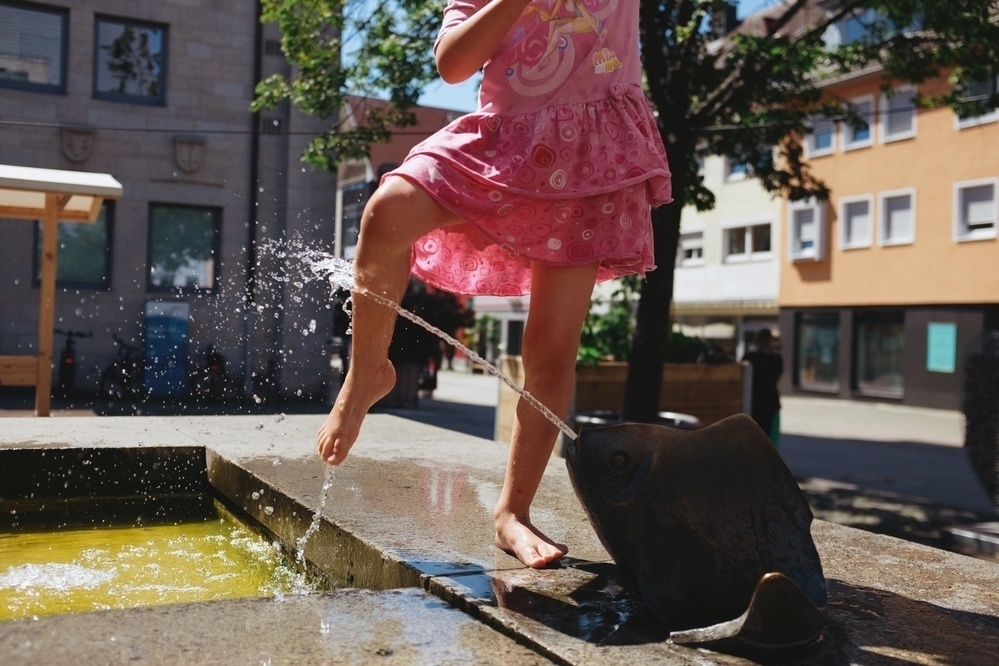 Young girl plays with water that shoots out of a decorative fish sculpture on a public city fountain on a hot summer day