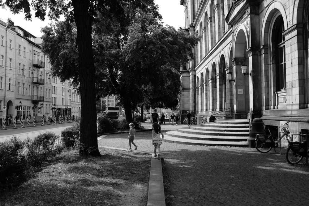 Black and white photograph of an outdoor urban scene with classical architecture, trees, a pedestrian path, and children playing.