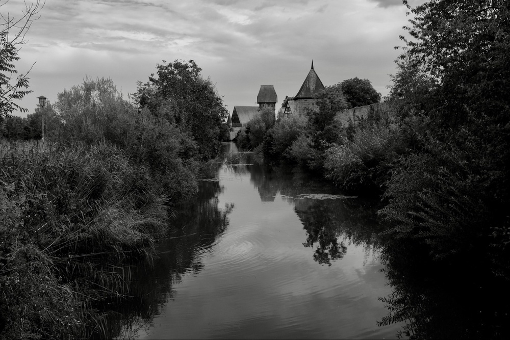 Black-and-white photo of the river Wörnitz reflecting the cloudy sky and sided by tall shrubs and trees. In the background, the centuries-old protection wall of Dinkelsbühl and two of its towers stand tall.