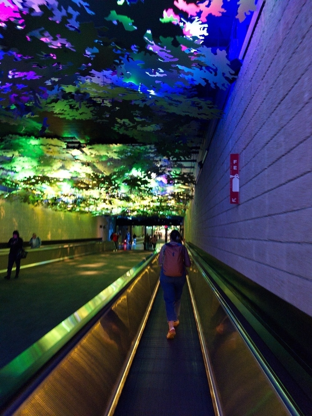 A person walks on a moving walkway beneath a ceiling illuminated with colorful, abstract lighting. Other individuals are visible in the background of the corridor, which has a modern and artistic design.