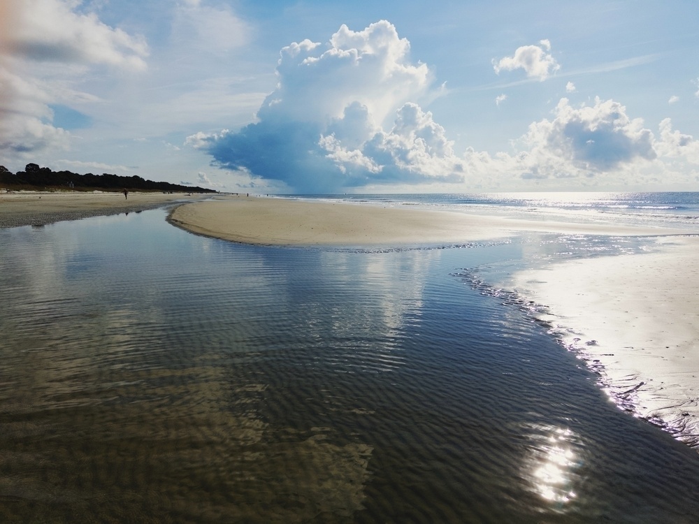 A serene beach scene with a partly cloudy sky. Calm water reflects the clouds above, and a narrow sandbar extends into the ocean. Distant figures walk along the shore with trees lining the horizon.