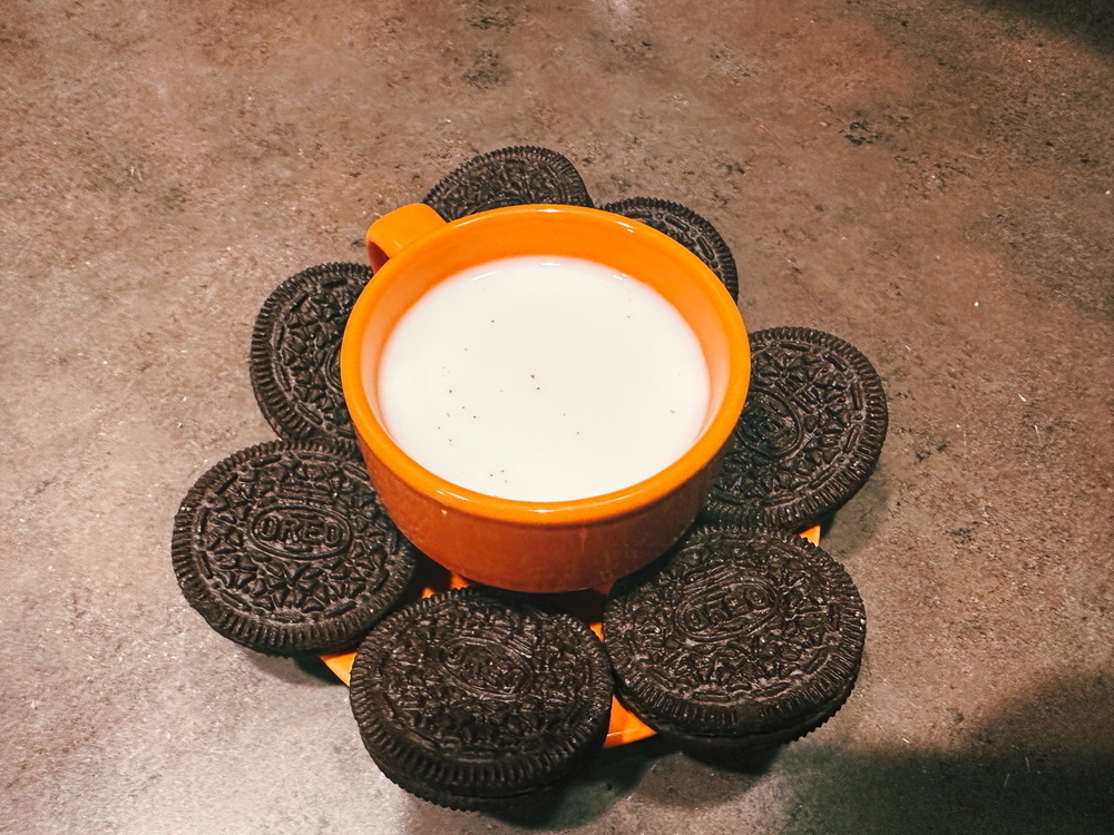 An orange cup filled with milk is placed on an orange plate. Surrounding the cup are several Oreo cookies arranged in a circular pattern on the plate. The setting is on a textured gray surface.