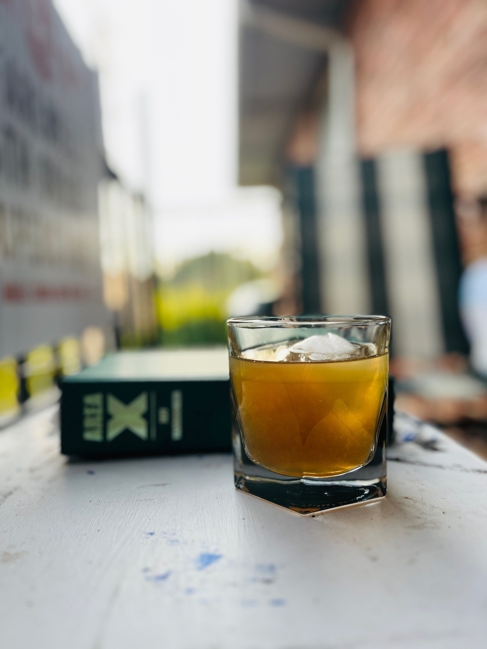 A whiskey glass with a cocktail in focus with a book behind it, sitting on a table, outdoors, background blurred