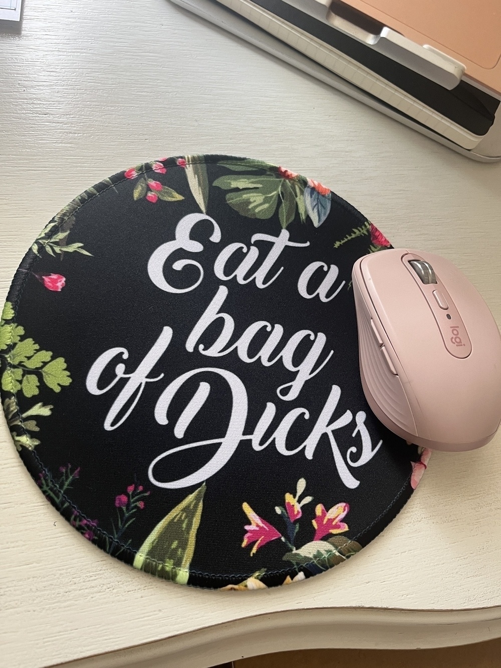 A black floral mousepad with the words “eat a bag of dicks” in script across it next to a pink computer mouse