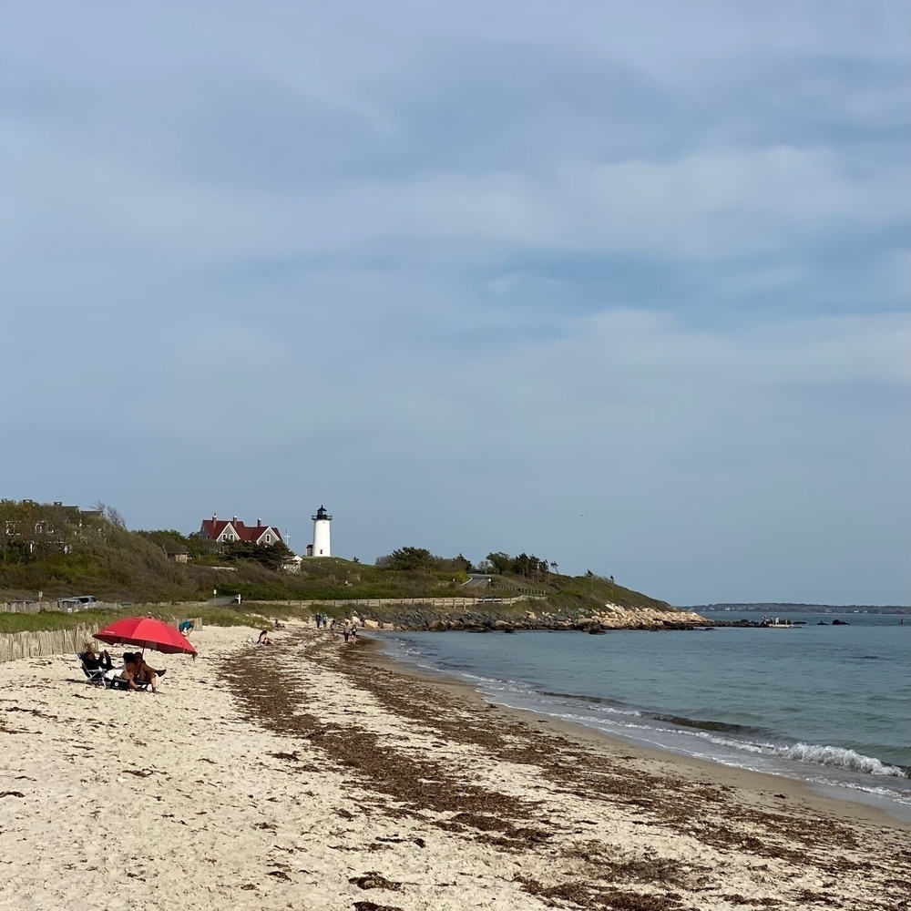 View of a beach with a white lighthouse on a point in the distance and a hazy sky above, a single red umbrella on the beach.