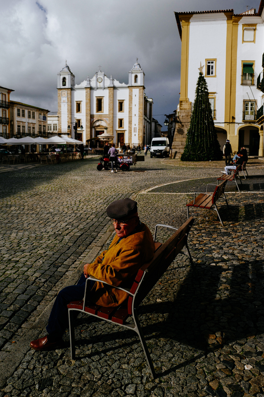 An old man sits on a bench, in a square. A church on the background. 