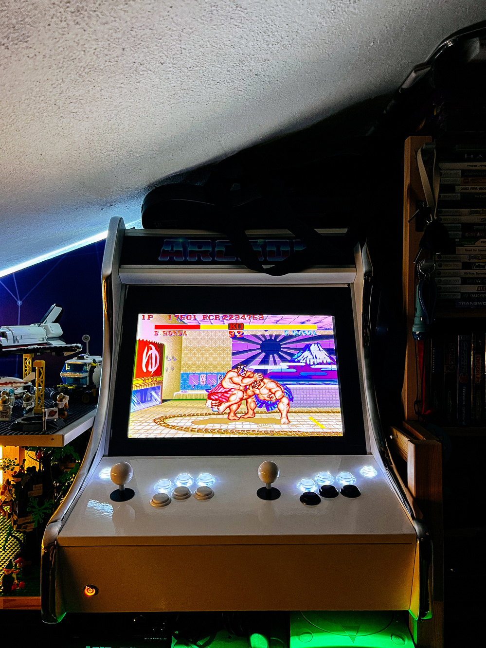 An arcade machine with the game &lsquo;Street Fighter II&rsquo; on the screen, set in a room with ambient lighting and shelves of books and Lego models.