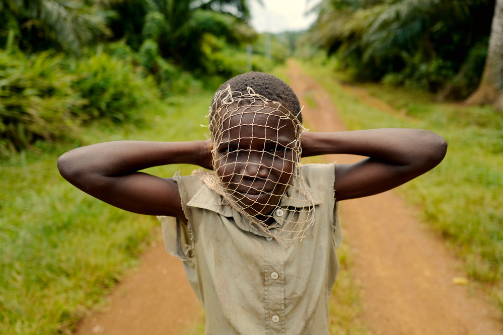 A kid looks at us, his hands holding a net to his face, like a mask.  Forest road in the background. 