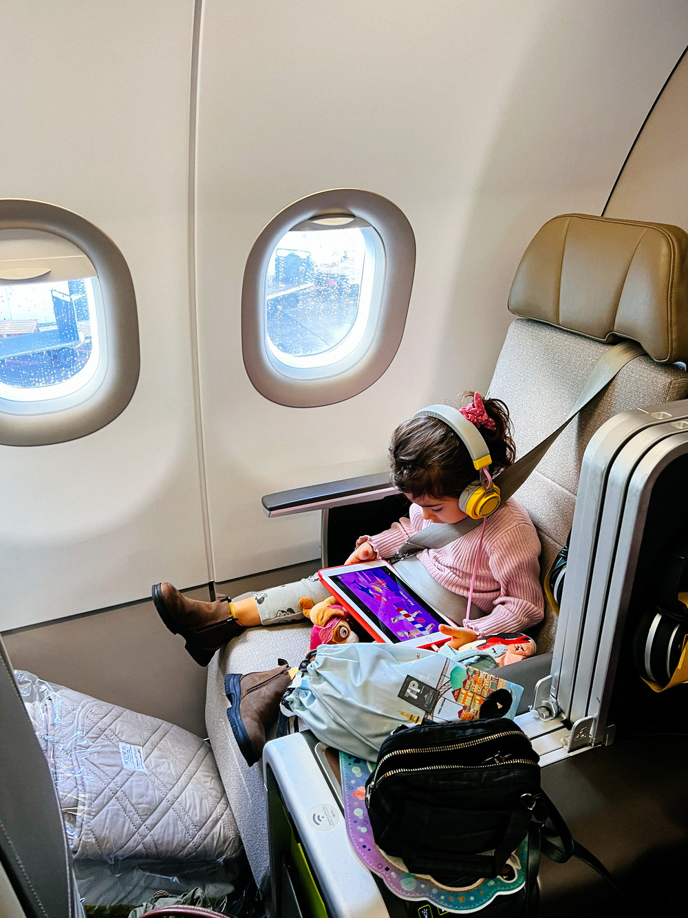 A toddler sits on an airplane seat, entertaining herself with an iPad. 