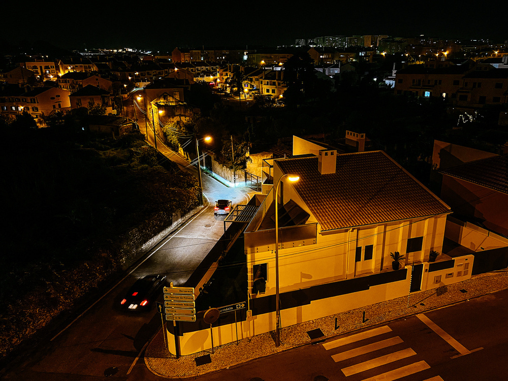 Nighttime view of a residential neighborhood with lit streetlights, houses with tiled roofs, and vehicles on the road; city lights in the background.
