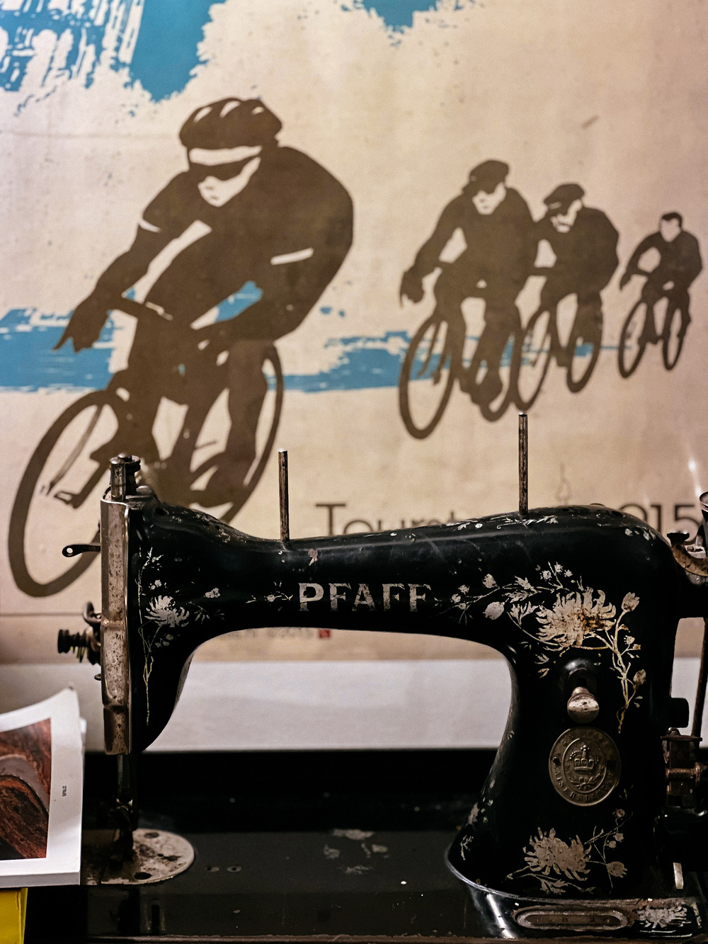 Vintage PFAFF sewing machine in the foreground with ornate decorations, placed against a backdrop of a large wall mural depicting cyclists in a race.