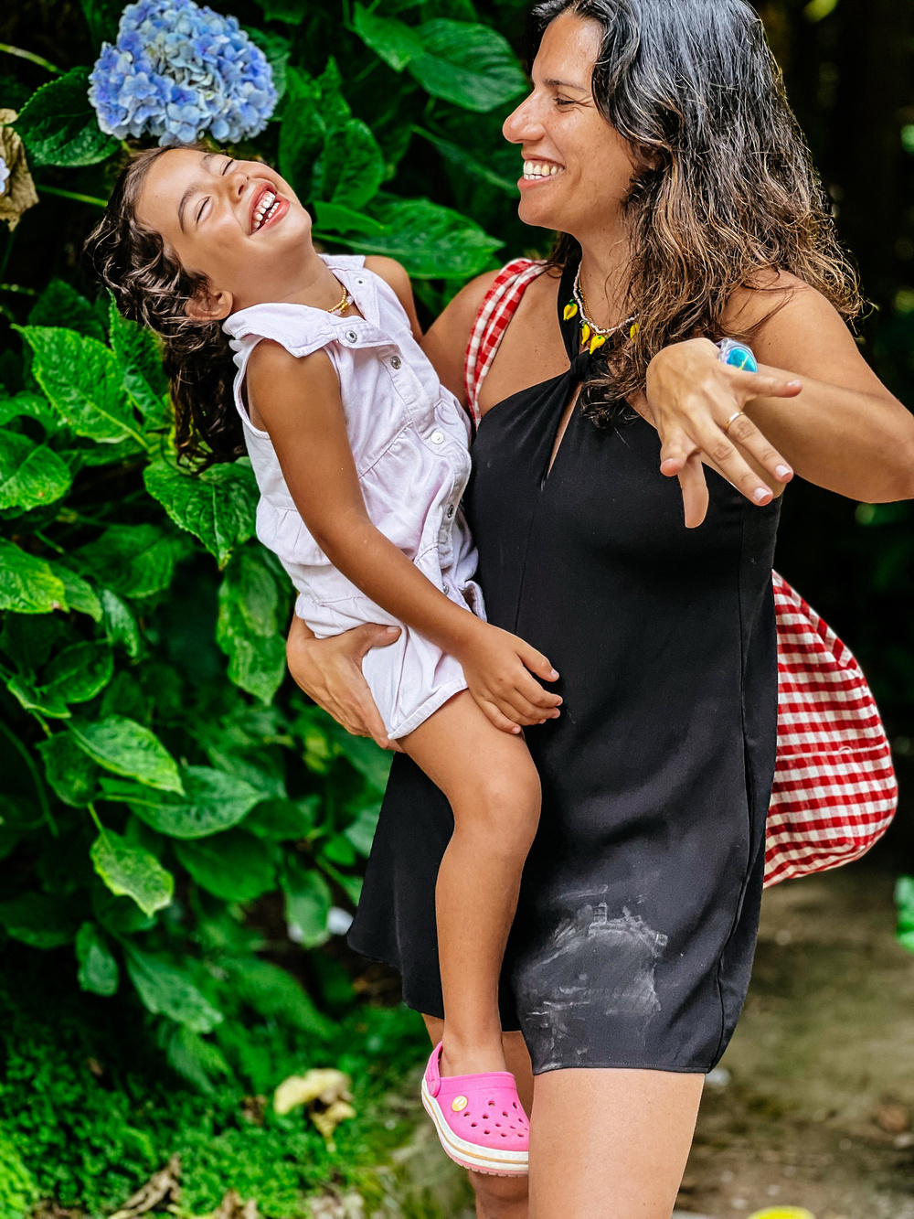 A woman in a black dress is holding a laughing child wearing a white dress and pink shoes. They are outdoors near green foliage and a blue hydrangea. The woman is smiling and has a checkered red and white bag on her shoulder.