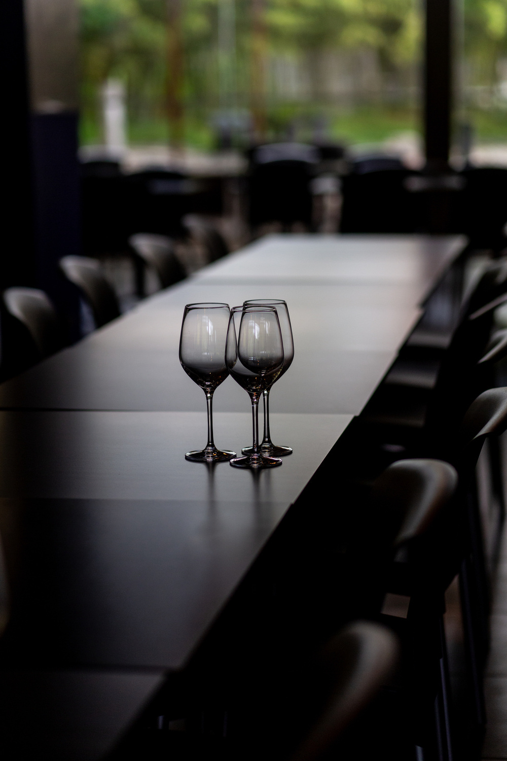 Three empty wine glasses on a long dark table with chairs lined up along it, with a blurred background of a room with large windows and outdoor greenery.