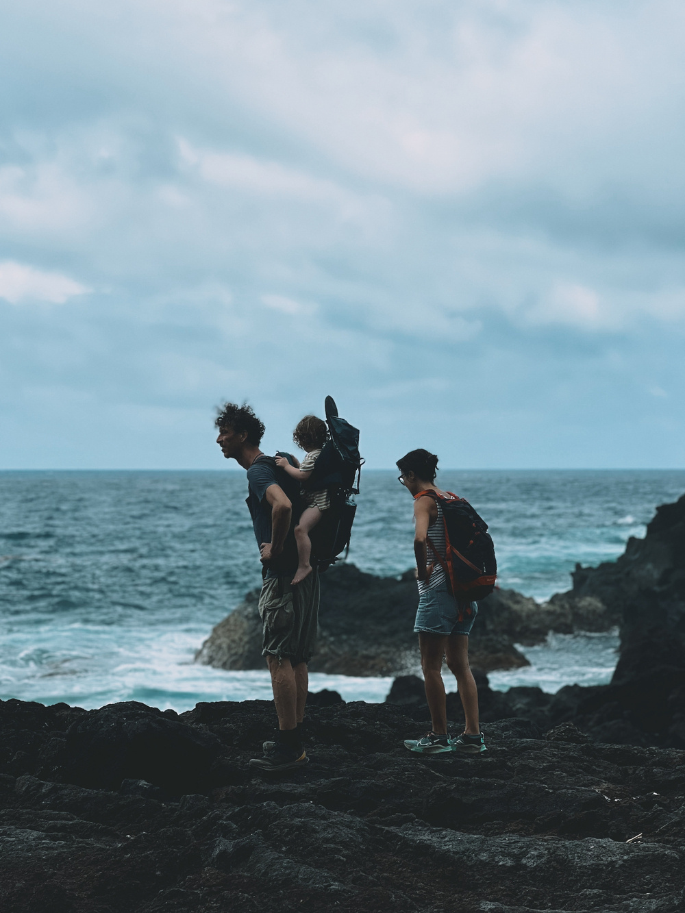 A family of three is shown hiking near a rocky coastline. An adult is carrying a young child in a backpack carrier, while another adult stands nearby with a backpack. The ocean waves crash against the rocks in the background under an overcast sky.