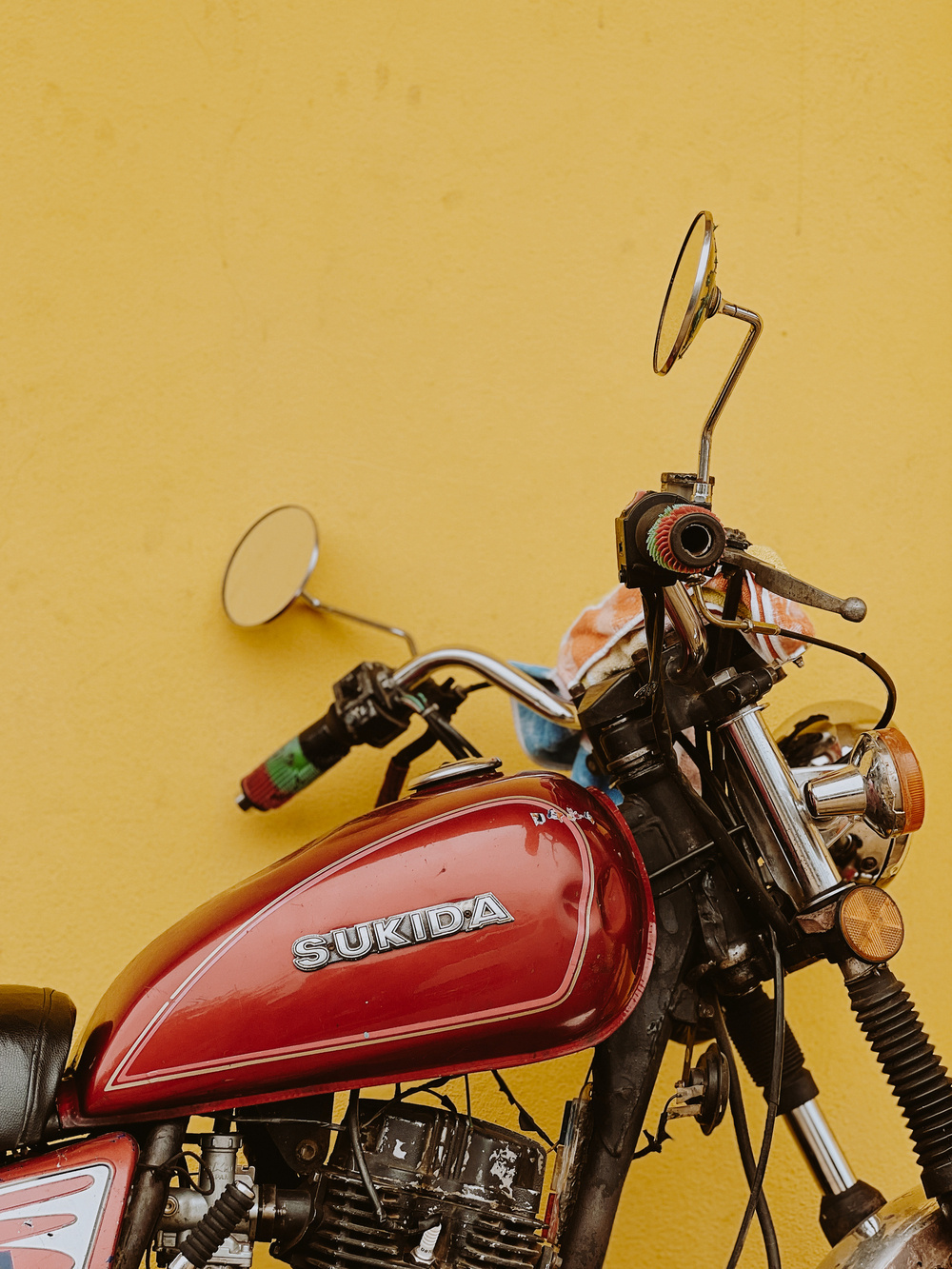 A close-up of a red Sukida motorcycle against a yellow wall. The motorcycle has rearview mirrors, a handlebar with colorful grips, and a front headlight. The image highlights the tank and part of the engine.