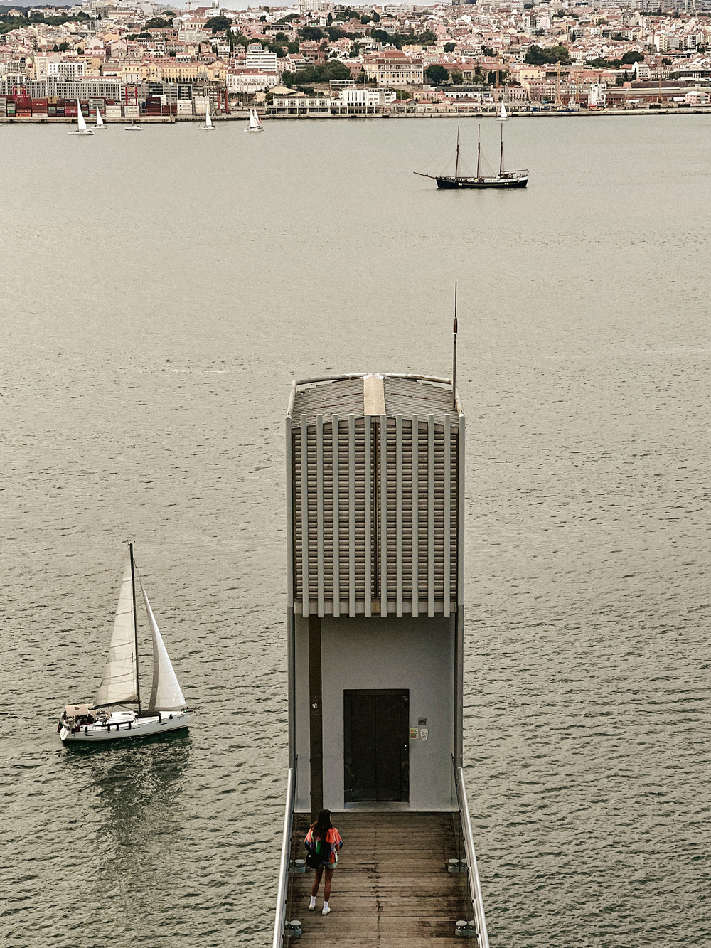 A person walks on a narrow wooden path leading to an elevator structure over a body of water. Sailboats and a large ship are visible in the water. In the background, there is a cityscape with numerous buildings and a port area.