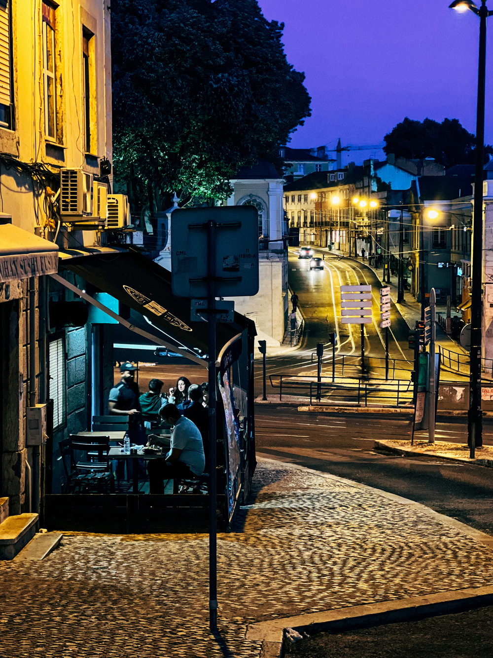 A nighttime street scene in a city, with a well-lit outdoor seating area where people are dining, adjacent to a cobblestone street with a few cars and illuminated streetlights.