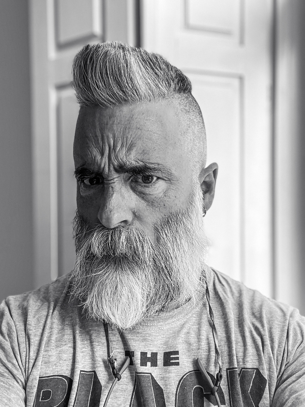 A black and white photo of a man with a styled pompadour haircut and a full beard, wearing a T-shirt and headphones around his neck. He appears to be indoors, with a door in the background.