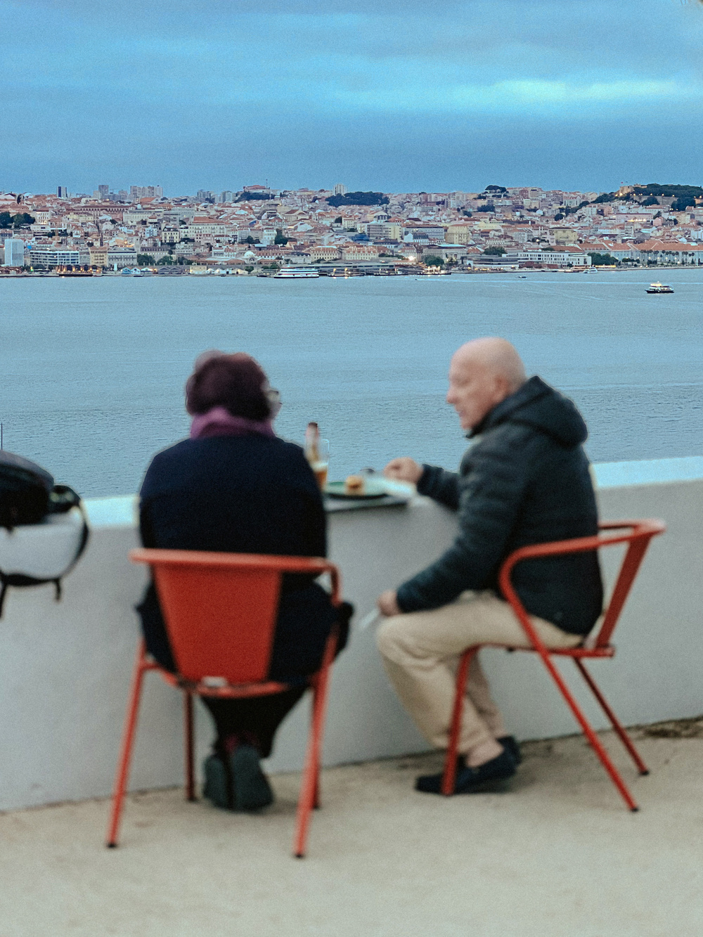 Two people sitting on red chairs at an outdoor terrace, overlooking a large body of water with a cityscape in the background. The people are engaged in conversation and have food items on the table. The weather appears to be cloudy.