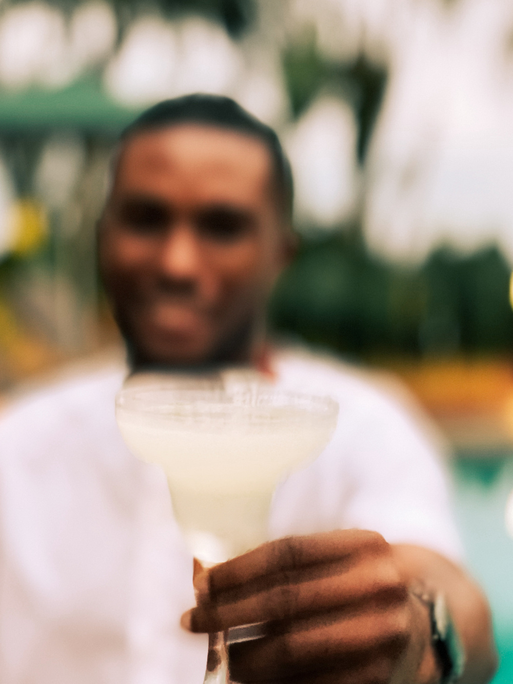 A person, with their face out of focus, is holding a clear glass with a light-colored drink. The background is an outdoor setting with greenery. 