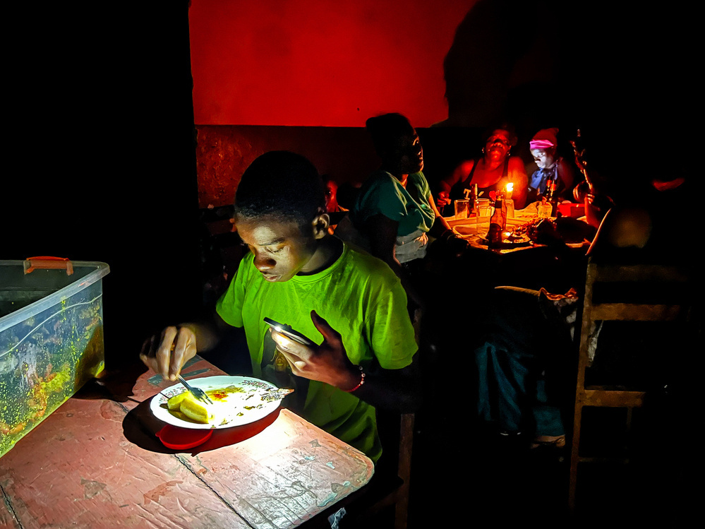 A boy in a green shirt eats food from a plate, illuminated by his phone’s light. In the background, a group of people sits around a candle-lit table, engaged in conversation. The scene is dimly lit with a strong red hue on