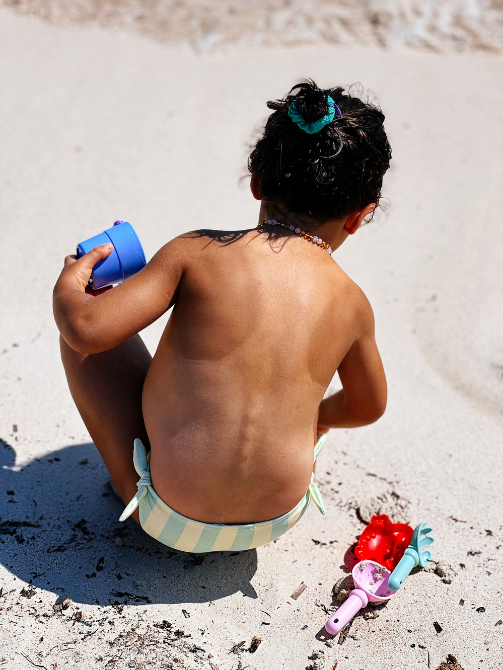 A young child is sitting in the sand at a beach, wearing a blue and white striped swimsuit bottom and a turquoise hair tie. The child is holding a blue toy cup and has a necklace on. There are sand toys, including a red crab mold.