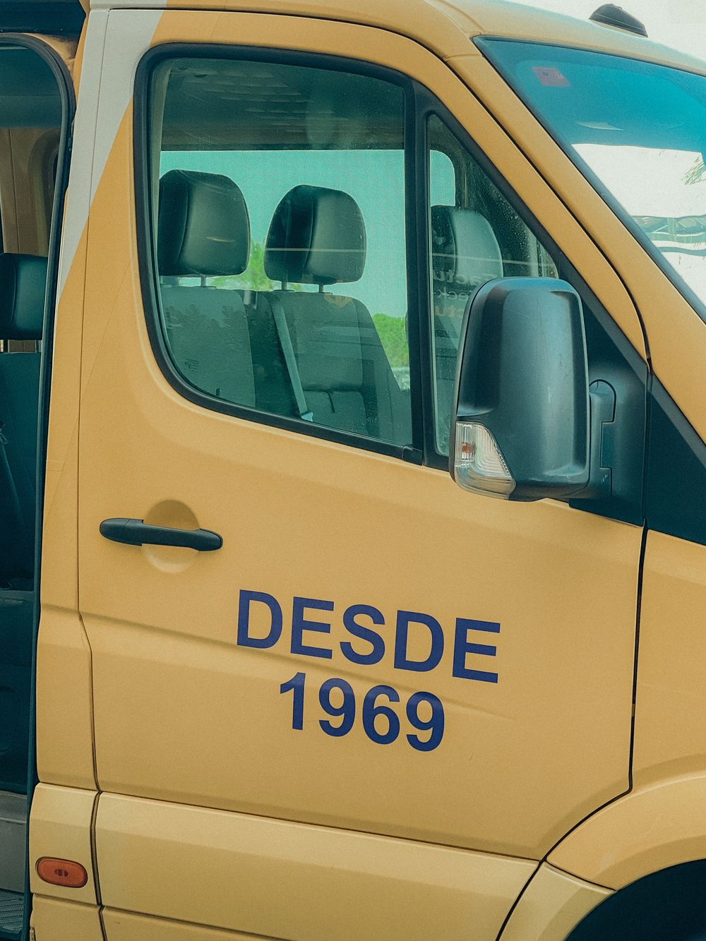 A partial view of a yellow van with the text &ldquo;DESDE 1969&rdquo; printed on the side. The focus is on the driver&rsquo;s side door and window.