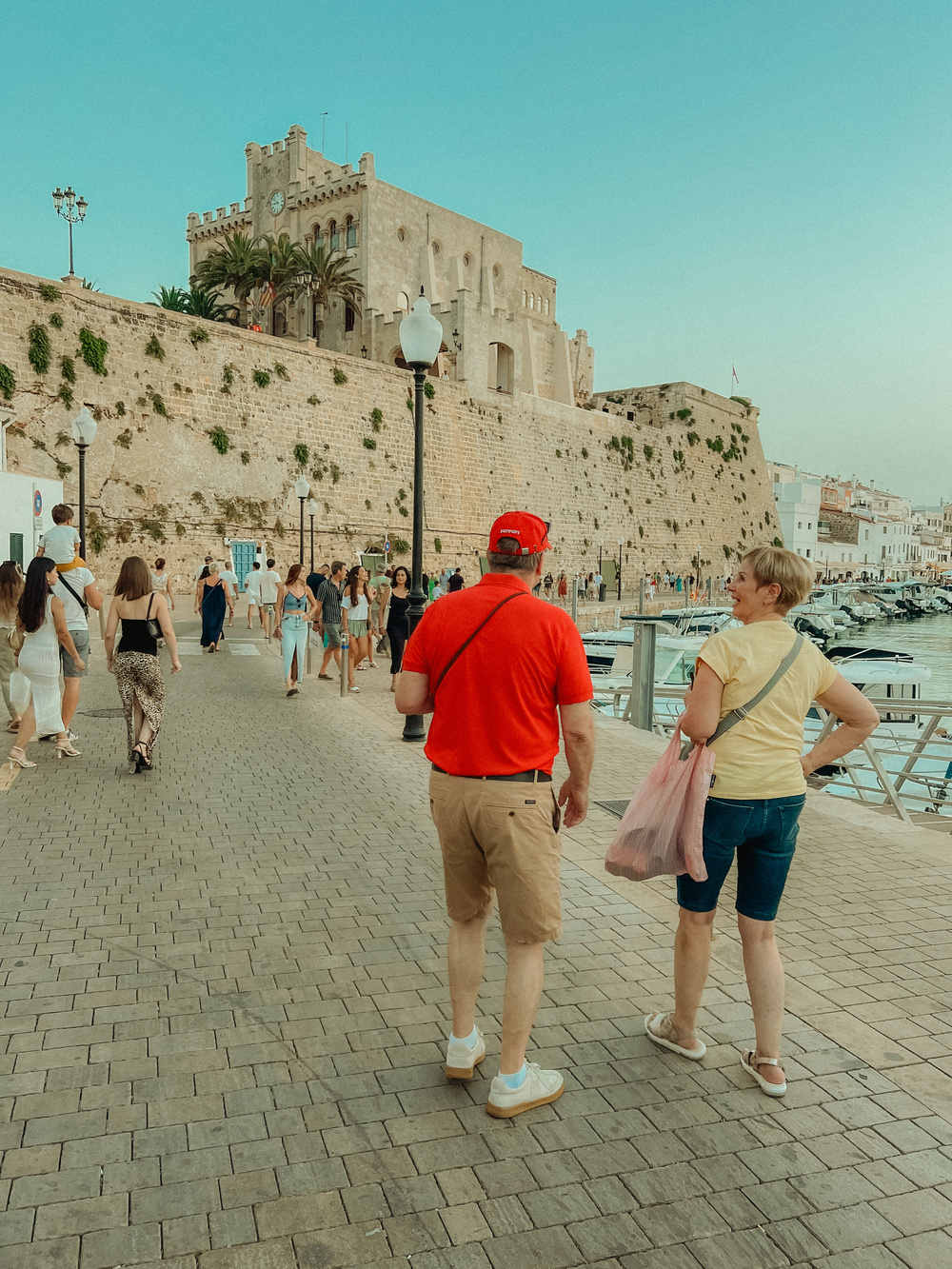 People walking on a cobblestone promenade next to a historic castle-like building. The sky is clear and blue. The scene includes several people, some near the stone wall and others by the water&rsquo;s edge where boats are docked.