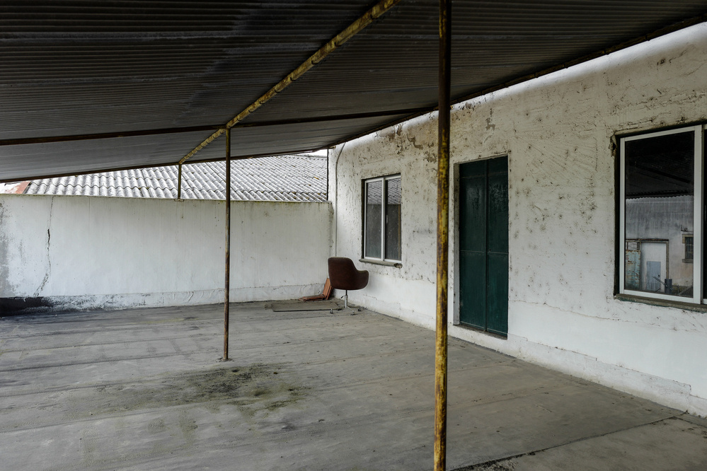 An outdoor area with a corrugated metal roof supported by rusted poles. The walls are white with visible wear and cracks. There are two windows and a green door. An old, brown chair is positioned near one wall. The concrete floor has stains.