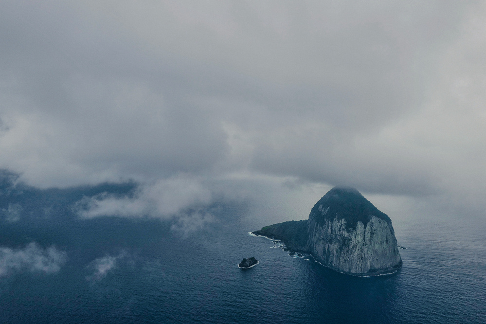 Aerial view of a large, dome-shaped rocky island in the ocean with a smaller rock formation nearby, under an overcast sky with low clouds.