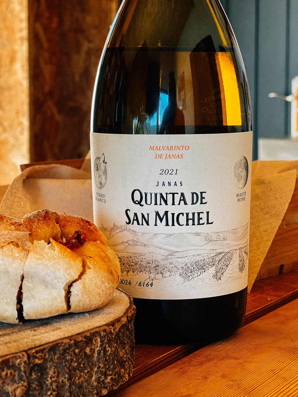 A bottle of white wine labeled “Malvarinto de Janas 2021 Quinta de San Michel” next to a round loaf of bread on a wooden table.