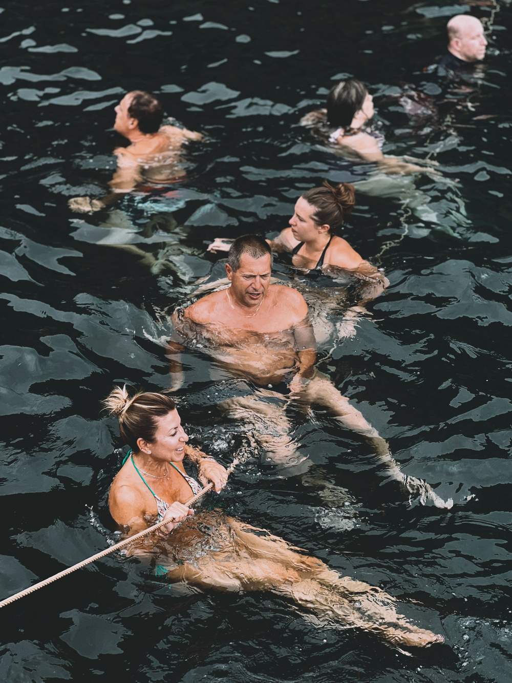 A group of people swimming in dark water. One person in the foreground is holding onto a rope, and the others are swimming nearby. Some are submerged more deeply than others.