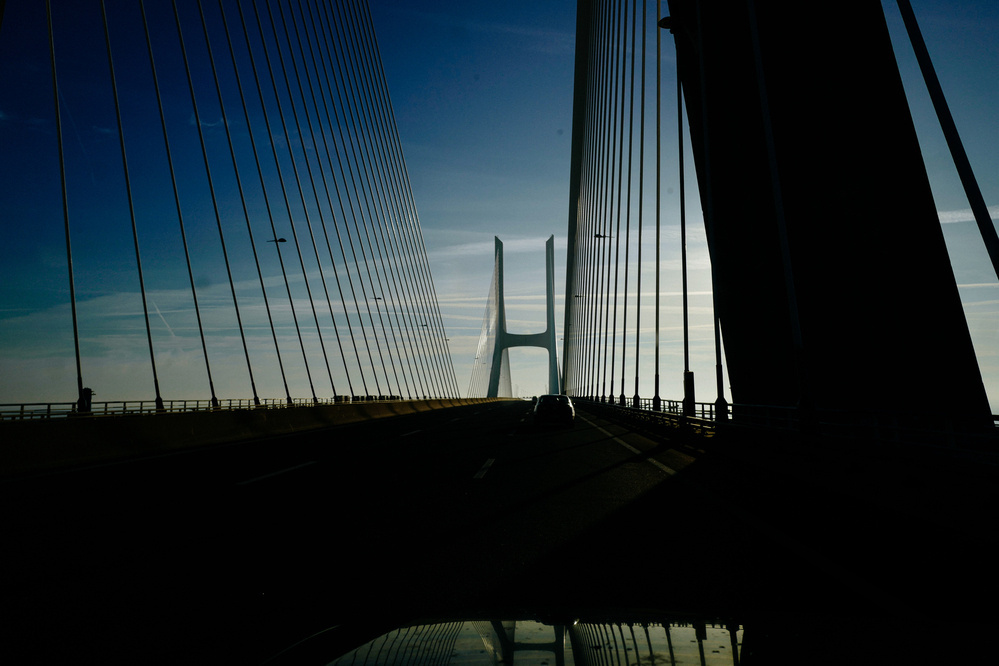 A silhouette of a cable-stayed bridge against a bright sky, with a car driving on the road and reflections visible on the vehicle&rsquo;s surface.