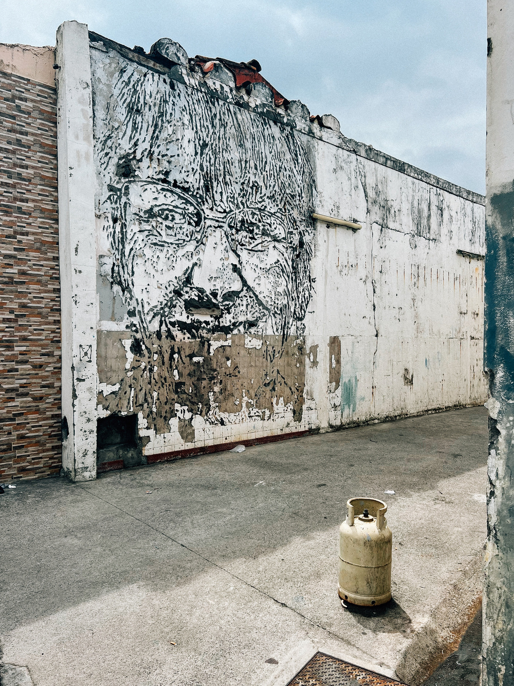 A dilapidated wall with a large, partially eroded mural of a face wearing glasses. The wall is adjacent to a building with textured brick-like tile. In the foreground, there is a single, old gas cylinder placed on the concrete pavement.