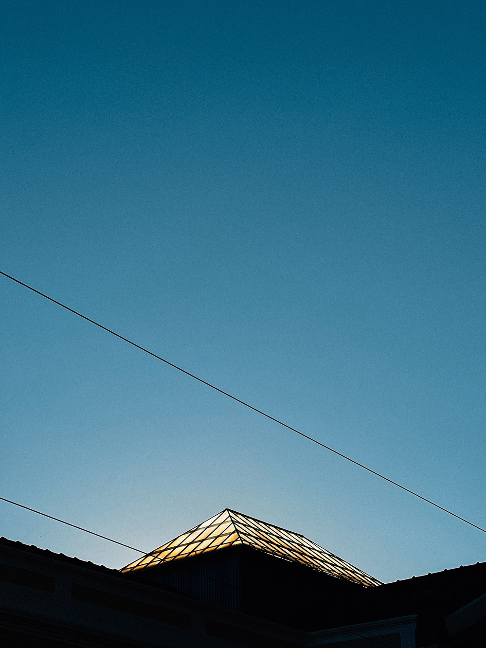 The image features the silhouette of a building with a glass skylight on its roof against a clear blue sky. Two overhead wires extend diagonally across the sky.