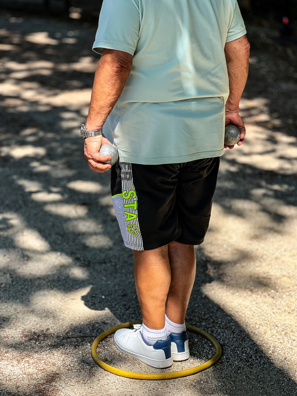 A person standing within a yellow circle on a gravel path, holding two metallic balls. The person is wearing a light-colored shirt, black shorts with green and gray stripes, white socks, and white sneakers.