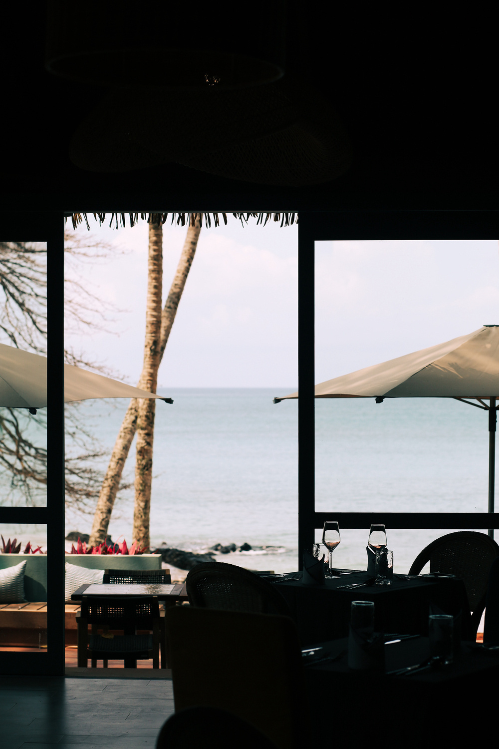 A beachfront restaurant with dark silhouetted tables and chairs in the foreground. Two wine glasses and napkins are set on one table. Through large windows, a view of the ocean, palm trees, and outdoor seating with umbrellas can be seen.