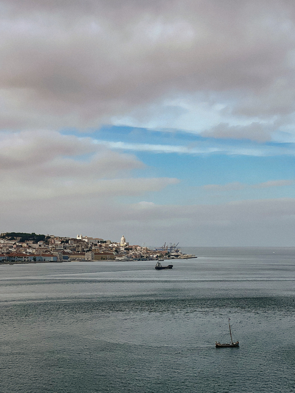 View of a coastal city with historic buildings and a clock tower, a calm body of water in the foreground with two boats, one near the shore and one closer to the camera, under a partly cloudy sky.