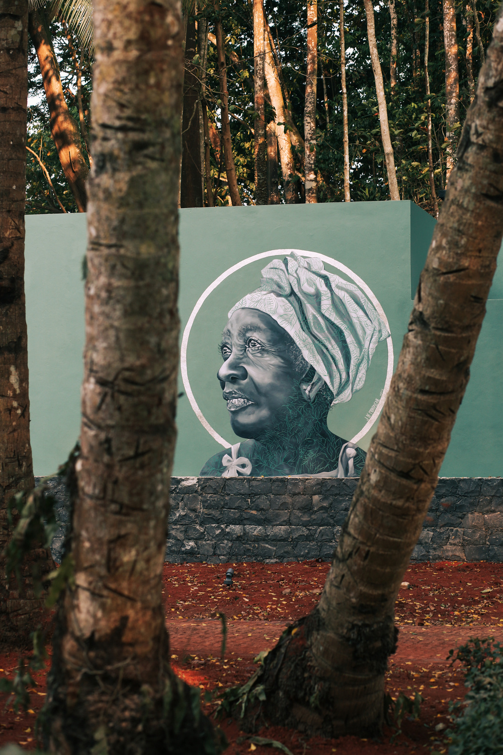 A mural painted on an outdoor wall features a side profile of an older woman wearing a headwrap. The mural is surrounded by a green circle and is partially obscured by tree trunks in the foreground.