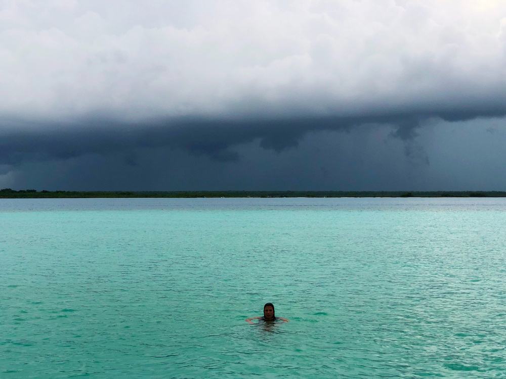 A person swimming in turquoise water with a backdrop of a dark, stormy sky.